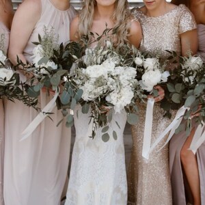 K&ouml;nnte beinhalten: F&uuml;nf Frauen in verschiedenen Schattierungen von Blush-Rosa und Goldkleidern halten Blumenstr&auml;u&szlig;e aus wei&szlig;en Blumen und Gr&uuml;n. Die Str&auml;u&szlig;e sind mit wei&szlig;em Satinband gebunden.
