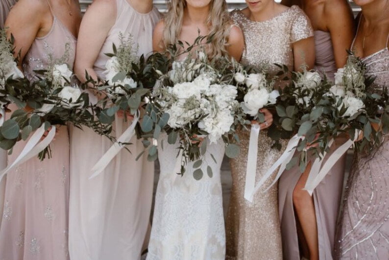 K&ouml;nnte beinhalten: F&uuml;nf Frauen in verschiedenen Schattierungen von Blush-Rosa und Goldkleidern halten Blumenstr&auml;u&szlig;e aus wei&szlig;en Blumen und Gr&uuml;n. Die Str&auml;u&szlig;e sind mit wei&szlig;em Satinband gebunden.