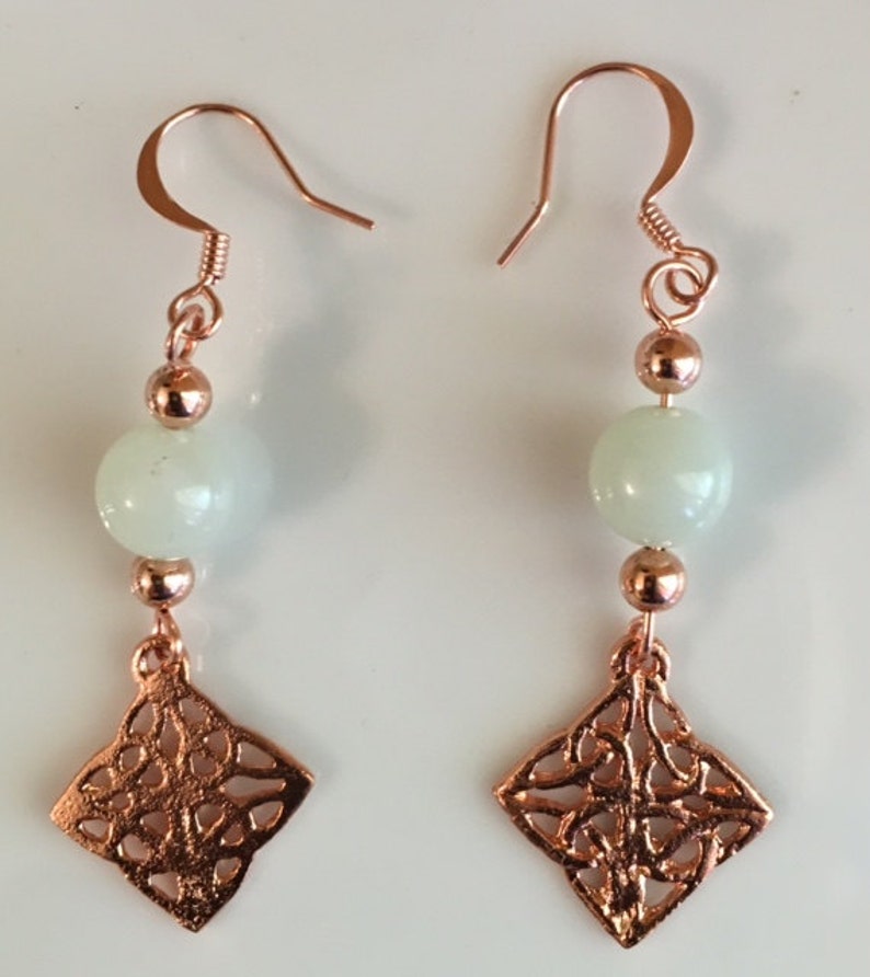 May include: A pair of copper earrings with a white stone bead and a copper filigree charm. The earrings have a hook closure.