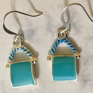 Pu&ograve; includere: Un paio di orecchini pendenti in argento con un design a corda blu e bianca e un ciondolo quadrato blu turchese.