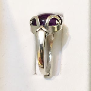 May include: A silver ring with a large, oval, deep purple gemstone. The ring has a decorative setting with curved and pointed elements. The band is smooth and polished, reflecting light. The ring is set against a white background.