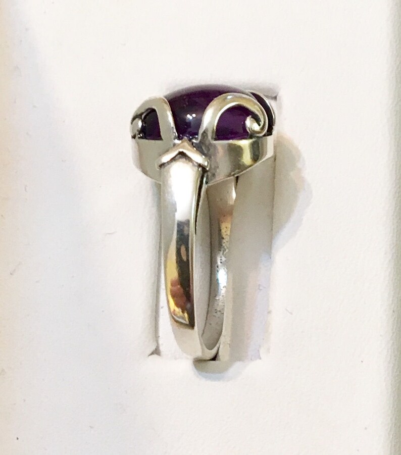 May include: A silver ring with a large, oval, deep purple gemstone. The ring has a decorative setting with curved and pointed elements. The band is smooth and polished, reflecting light. The ring is set against a white background.