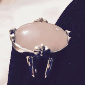 May include: A silver ring with a large, oval-shaped, pale pink gemstone set in a decorative, ornate setting.