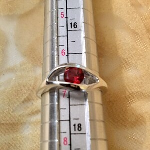 May include: A silver ring with a red gemstone set in a unique design. The ring is sized on a ring sizer, showing a size 7.