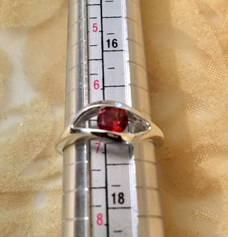 May include: A silver ring with a red gemstone set in a unique design. The ring is sized on a ring sizer, showing a size 7.