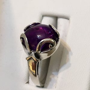 May include: A silver ring featuring a large, round, deep purple gemstone. The gemstone is held in place by decorative silver prongs with a swirling design. The ring has a polished, reflective finish.