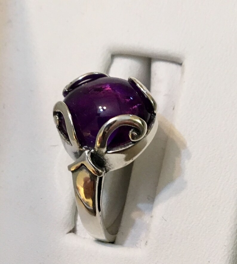 May include: A silver ring featuring a large, round, deep purple gemstone. The gemstone is held in place by decorative silver prongs with a swirling design. The ring has a polished, reflective finish.