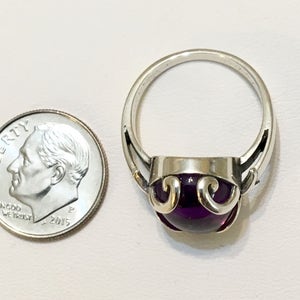May include: A silver ring with a large, round, deep purple gemstone. The ring has a decorative design around the gemstone. A U.S. dime is next to the ring for size comparison.