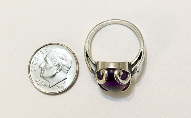 May include: A silver ring with a large, round, deep purple gemstone. The ring has a decorative design around the gemstone. A U.S. dime is next to the ring for size comparison.