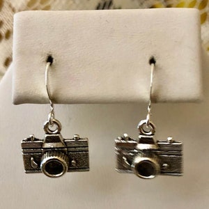 May include: A pair of silver camera dangle earrings. The camera charms are detailed with a lens and a textured surface.