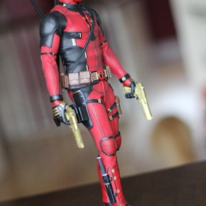 Peut inclure: Une figurine d'action rouge et noire de Deadpool tenant deux pistolets dorés. La figurine porte une combinaison rouge et noire avec une ceinture noire et un holster noir sur la jambe droite. La figurine est debout sur une surface brun foncé.