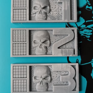 Puede incluir: Tres fichas de plástico gris con un diseño de calavera y placa de circuito. Cada ficha tiene un número, 1, 2 y 3, impreso en ella.
