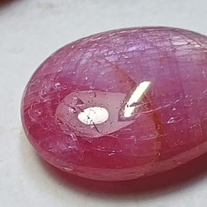Peut inclure: Une pierre précieuse polie, de forme ovale, dans des tons roses et rouges. La surface présente un éclat brillant avec des inclusions internes et un reflet. La gemme est probablement un rubis, pour bijoux ou collection.