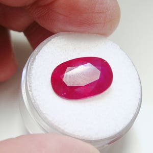 May include: A vibrant red oval-shaped gemstone in a white display container.