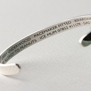 Puede incluir: Una pulsera de plata con texto grabado. El texto incluye un nombre, información médica y datos de contacto. La pulsera es curvada y abierta, diseñada para ser usada en la muñeca.