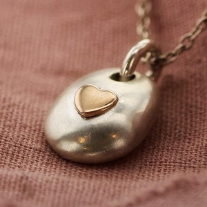 May include: A silver necklace with a pendant featuring a gold heart. The pendant is an oval shape with a small gold heart in the center. The necklace is displayed on a pink fabric background.