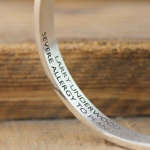 May include: A silver-colored bangle bracelet with engraved text. The text reads "SEVERE ALLERGY TO PEANUTS" and "LARRY UNDERWOOD". The bracelet is curved and reflects light, set against a blurred wooden background.