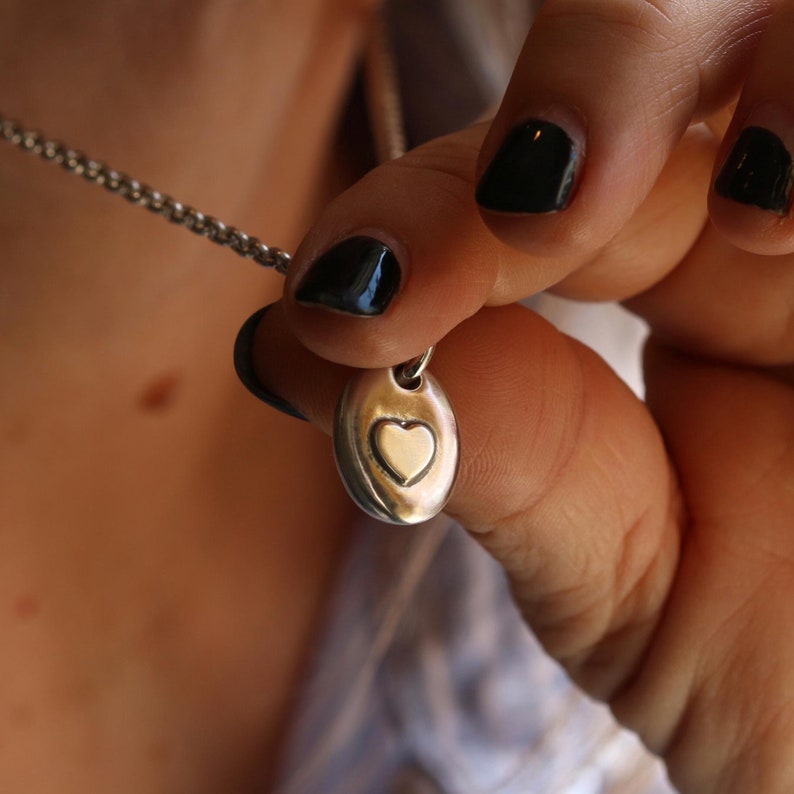 May include: Silver-toned necklace with a small, oval pendant. The pendant features a heart-shaped design in the center. The necklace is held by a person with dark nail polish. The chain is a delicate, linked style.