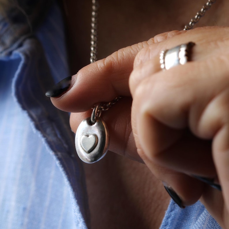 May include: A silver pendant necklace featuring an oval-shaped pendant with a heart design. The necklace is held by a person wearing a blue shirt. A silver ring is also visible.