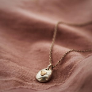 May include: A silver-toned pendant necklace with a small gold heart-shaped accent. The pendant has a smooth, organic shape and hangs from a delicate chain. The background is a soft, muted pink fabric.