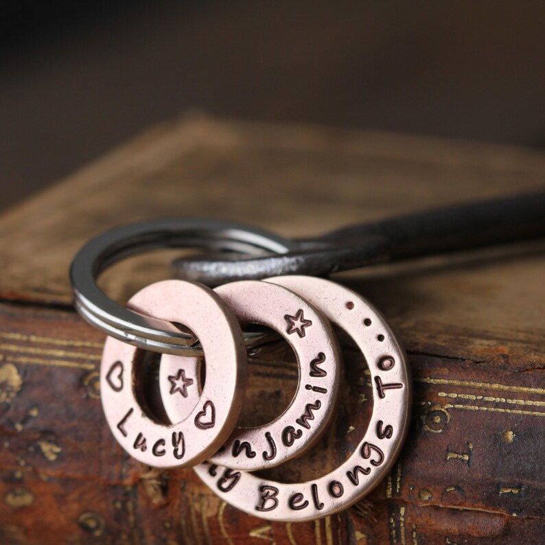 May include: A set of four silver-toned metal keychains with engraved text. The keychains are in the shape of rings with the words "Lucy", "My", "Jamil", and "Belongs To" engraved on them. The keychains are attached to a silver-toned metal keyring.