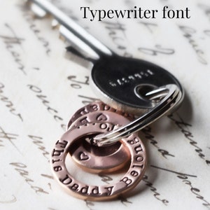 May include: Close-up of a silver key chain with three copper rings. The rings have the words "This Daddy" and "Belongs" engraved on them in a typewriter font.