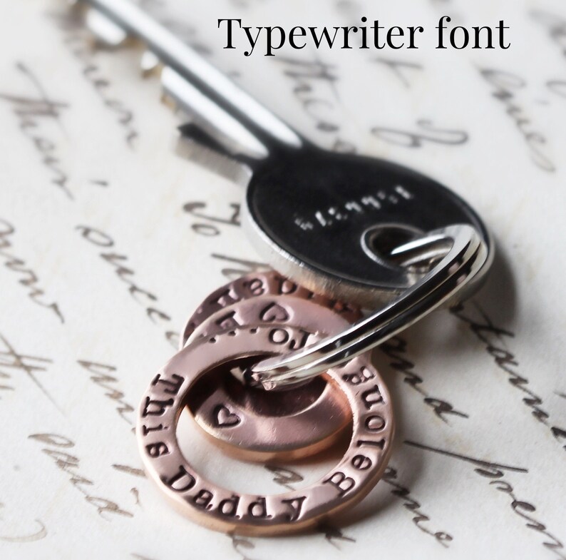 May include: Close-up of a silver key chain with three copper rings. The rings have the words "This Daddy" and "Belongs" engraved on them in a typewriter font.