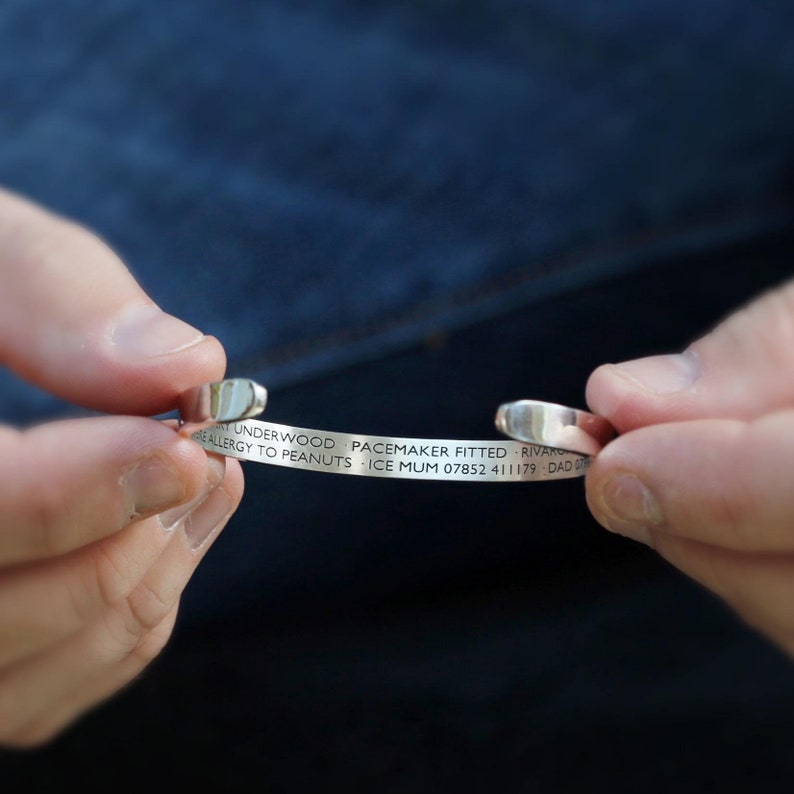 May include: A silver-colored medical alert cuff bracelet held by a person. The bracelet has engraved text including a name, medical condition, and emergency contact information. The bracelet is open-ended and has rounded edges.