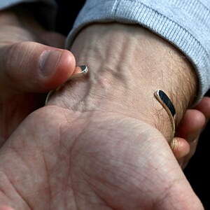 May include: A silver cuff bracelet with a minimalist design. The bracelet has a curved shape and is open at the ends. The ends of the bracelet are flat and have a dark, possibly black, inlay. The bracelet is being worn on a wrist.