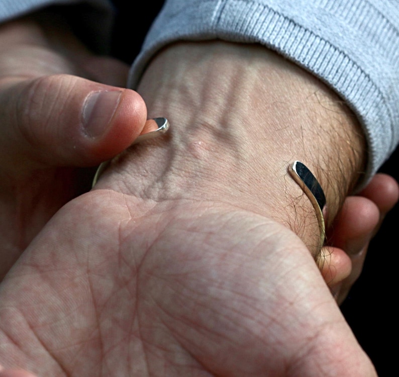 May include: A silver cuff bracelet with a minimalist design. The bracelet has a curved shape and is open at the ends. The ends of the bracelet are flat and have a dark, possibly black, inlay. The bracelet is being worn on a wrist.