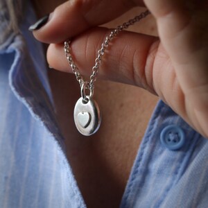 May include: A silver pendant necklace featuring an oval-shaped charm with a heart design. The pendant hangs from a delicate chain. The necklace is held up against a blue shirt.