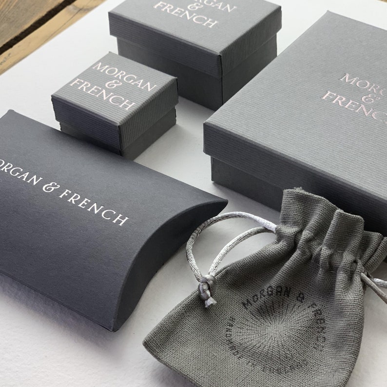 May include: An assortment of gray gift boxes and a drawstring pouch. The boxes are various sizes, some with textured surfaces, and all feature the text "MORGAN & FRENCH" in a contrasting color. The pouch also displays the brand name.