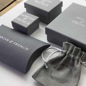 May include: An assortment of gray jewelry boxes and a drawstring pouch. The boxes are various sizes and feature the text "MORGAN & FRENCH" in white or rose gold. The pouch also has the brand name and the text "HANDMADE IN ENGLAND".