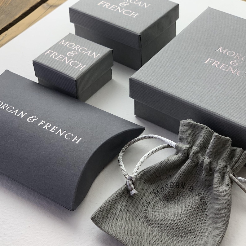 May include: An assortment of gray jewelry boxes and a drawstring pouch. The boxes are various sizes and feature the text "MORGAN & FRENCH" in white or rose gold. The pouch also has the brand name and the text "HANDMADE IN ENGLAND".
