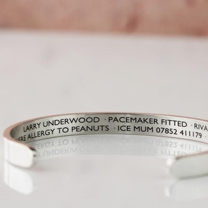 May include: A silver-colored cuff bracelet with engraved text. The text includes the name "LARRY UNDERWOOD", medical information, and a phone number. The bracelet is reflective and sits on a reflective surface.