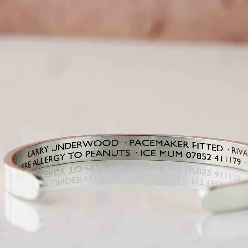 May include: A silver-colored cuff bracelet with engraved text. The text includes the name "LARRY UNDERWOOD", medical information, and a phone number. The bracelet is reflective and sits on a reflective surface.