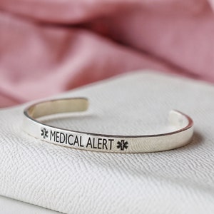 May include: A silver-colored medical alert cuff bracelet. The bracelet has the words "MEDICAL ALERT" engraved in black, along with a medical symbol on each side. The bracelet is open-ended and sits on a white surface.
