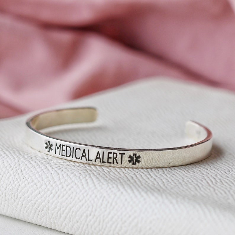 May include: A silver-colored medical alert cuff bracelet. The bracelet has the words "MEDICAL ALERT" engraved in black, along with a medical symbol on each side. The bracelet is open-ended and sits on a white surface.