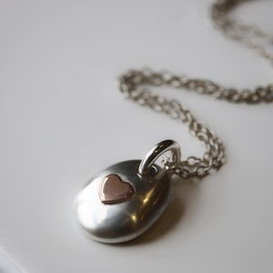 May include: A silver pendant necklace with a small rose gold heart. The pendant is oval-shaped and hangs from a delicate silver chain. The necklace is displayed on a white surface.