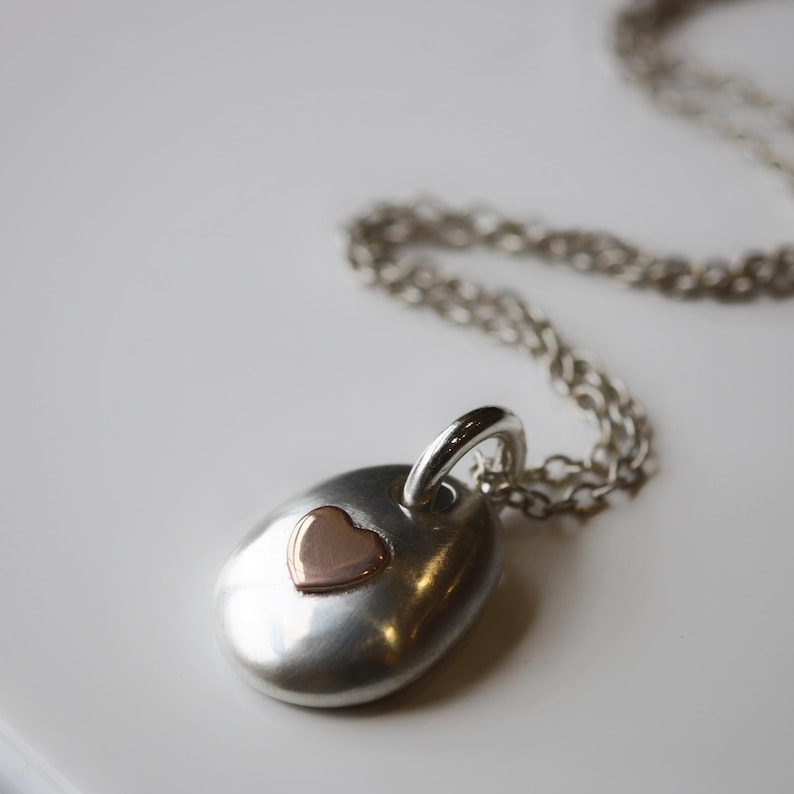 May include: A silver pendant necklace with a small rose gold heart. The pendant is oval-shaped and hangs from a delicate silver chain. The necklace is displayed on a white surface.