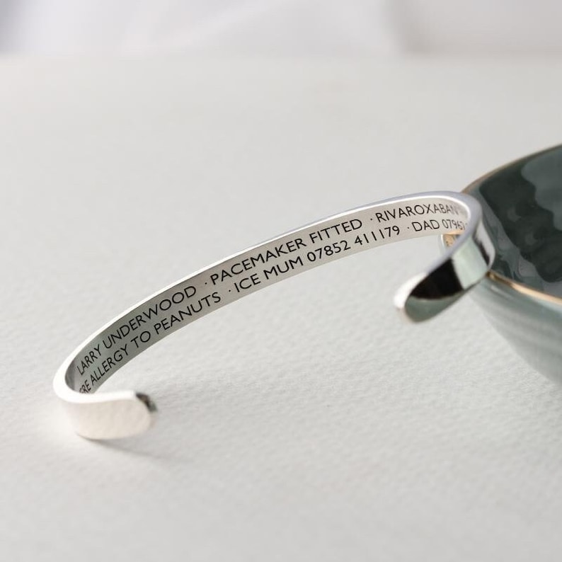 May include: A silver-colored cuff bracelet with engraved text. The text includes a name, medical information, and contact details. The bracelet is curved and open-ended, designed to be worn on the wrist.