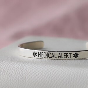 May include: A silver-colored medical alert cuff bracelet. The bracelet has the words "MEDICAL ALERT" in black lettering, along with the medical alert symbol on each side. The bracelet is resting on a white surface with a pink background.