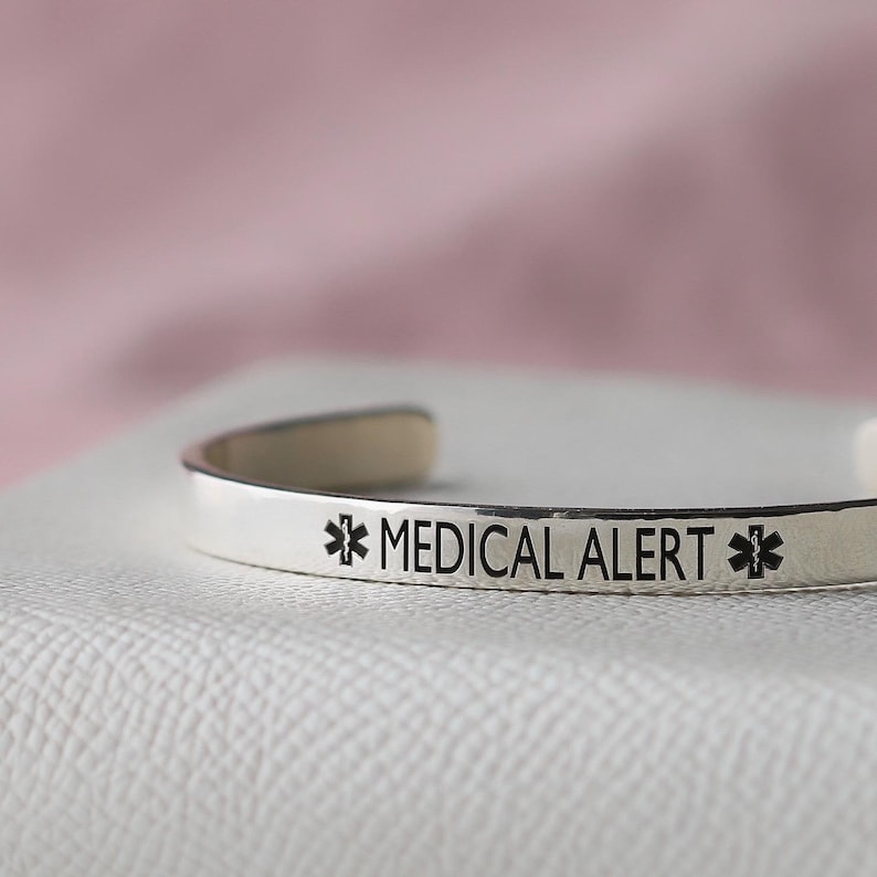 May include: A silver-colored medical alert cuff bracelet. The bracelet has the words "MEDICAL ALERT" in black lettering, along with the medical alert symbol on each side. The bracelet is resting on a white surface with a pink background.