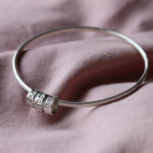 May include: A silver bangle bracelet with three silver rings attached. The rings have the words "DURA", "DON", and "SER" engraved on them.