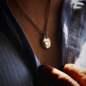 May include: Silver-toned necklace with a pendant featuring a heart-shaped design. The pendant is oval-shaped with a smaller heart in the center. The necklace is worn over a blue button-down shirt.