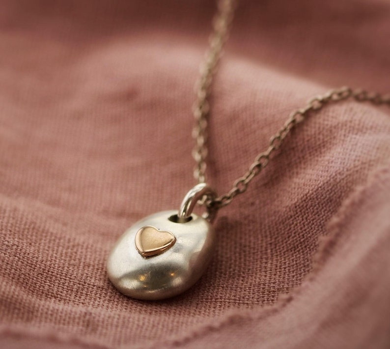 May include: A silver necklace with a pendant featuring a gold heart. The pendant is an oval shape with a small gold heart in the center. The necklace is displayed on a pink fabric background.