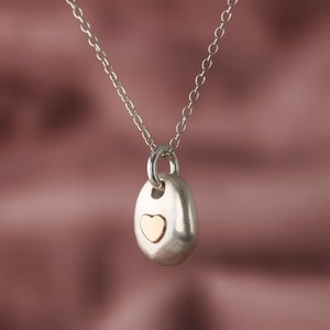May include: A silver pendant necklace featuring a smooth, oval-shaped pendant with a gold heart inset. The pendant hangs from a delicate silver chain. The background is a soft, muted pink.