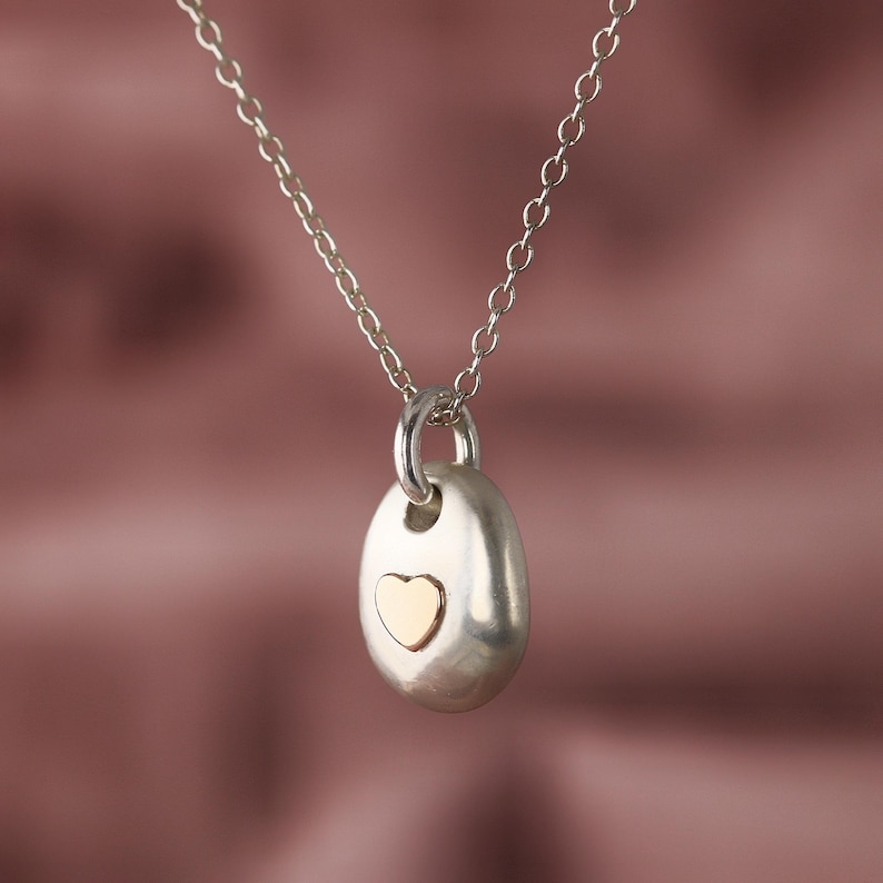 May include: A silver pendant necklace featuring a smooth, oval-shaped pendant with a gold heart inset. The pendant hangs from a delicate silver chain. The background is a soft, muted pink.