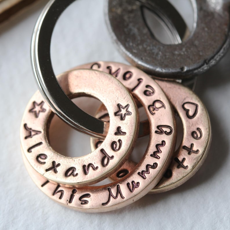 May include: A set of five keychains with a silver ring. The keychains are made of copper and have engraved words on them. The words are "This", "Alexander", "Mummy", "Love", and "Little".