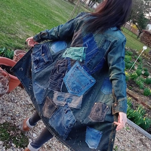 Op de afbeelding: Lange spijkerjas met meerdere zakken en patches gemaakt van gerecyclede jeans. De jas is een patchwork van verschillende tinten blauwe denim, waardoor een unieke en getextureerde uitstraling ontstaat. De jas wordt buiten gedragen.